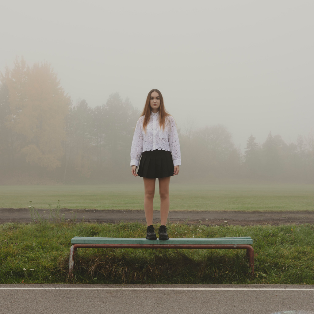 Photo by Samuel Galanský: Girl in a white shirt and black skirt standing on a bench outside in foggy weather, facing the camera.