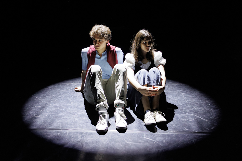 Photo by Samuel Galanský: Two young people sitting cross-legged on a dark stage, illuminated by a single spotlight.